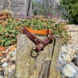 A brown and orange dog collar with gold rings is draped over a weathered wooden post in a garden with colourful flowers, gravel, and a wooden fence under a partly cloudy sky.