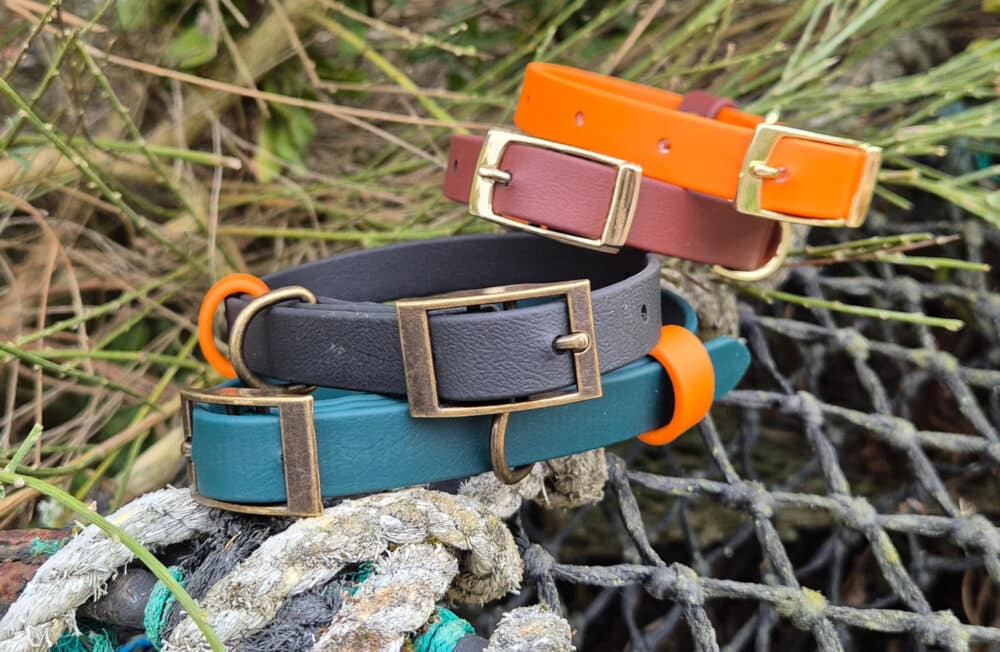 Four handmade Biothane dog collars in teal, black, maroon, and orange with brass buckles are displayed on a fishing net and rope, with greenery in the background.