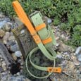 The Bordeaux Grove Extra Wide Collar and matching green and orange lead set, crafted from sturdy material, is displayed on rocks beside a wooden post with green leafy plants in the background.
