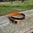Handmade Amberwood Biothane dog collar with orange and brown BioThane straps and brass buckle, displayed on rustic wooden table overlooking pastoral countryside landscape.