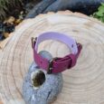 A pink and purple buckle bracelet is displayed on a grey stone with a hole, placed on a tree stump outdoors. Green plants and a rabbit garden ornament are visible in the background.