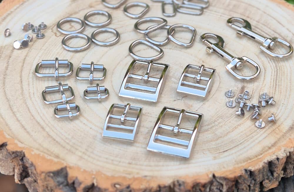 Hand-crafted stainless steel hardware components for dog collars displayed on wood, including D-rings, buckles and fasteners.