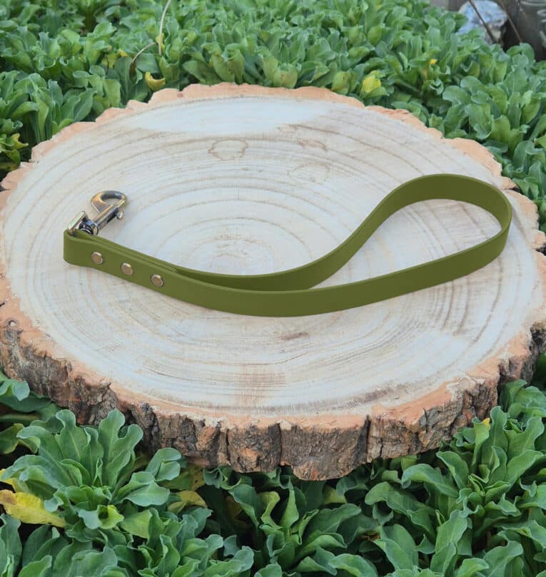 A Custom Single Colour Traffic Handle in olive green with a metal clasp rests on a round wooden tree stump, surrounded by green leafy plants.