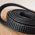 A close-up of the Super-grip Biothane Training Tab / Obedience Lead 51cm, a black textured strip with raised circular bumps, resting on wood. Flexible and rubbery, it's ideal for grip or as a protective covering.