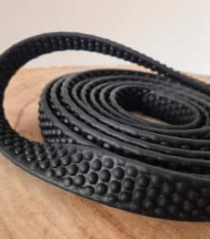 A close-up of the Super-grip Biothane Training Tab / Obedience Lead 51cm, a black textured strip with raised circular bumps, resting on wood. Flexible and rubbery, it's ideal for grip or as a protective covering.