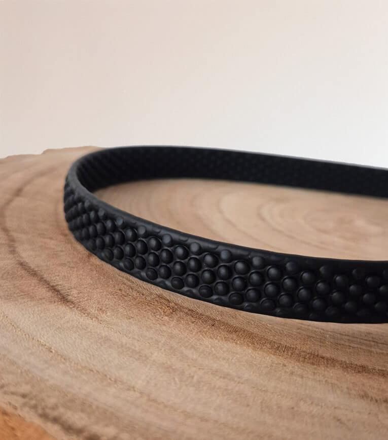 A black, textured Super-grip Biothane Training Tab / Obedience Lead 51cm with a raised dot pattern rests on a smooth, light wooden surface against a softly lit, plain background.