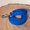 A coiled bright blue Waterproof Hexa Long-Line / Training Lead with a metal clasp and three silver rivets, shown on a round wooden surface with visible tree rings.