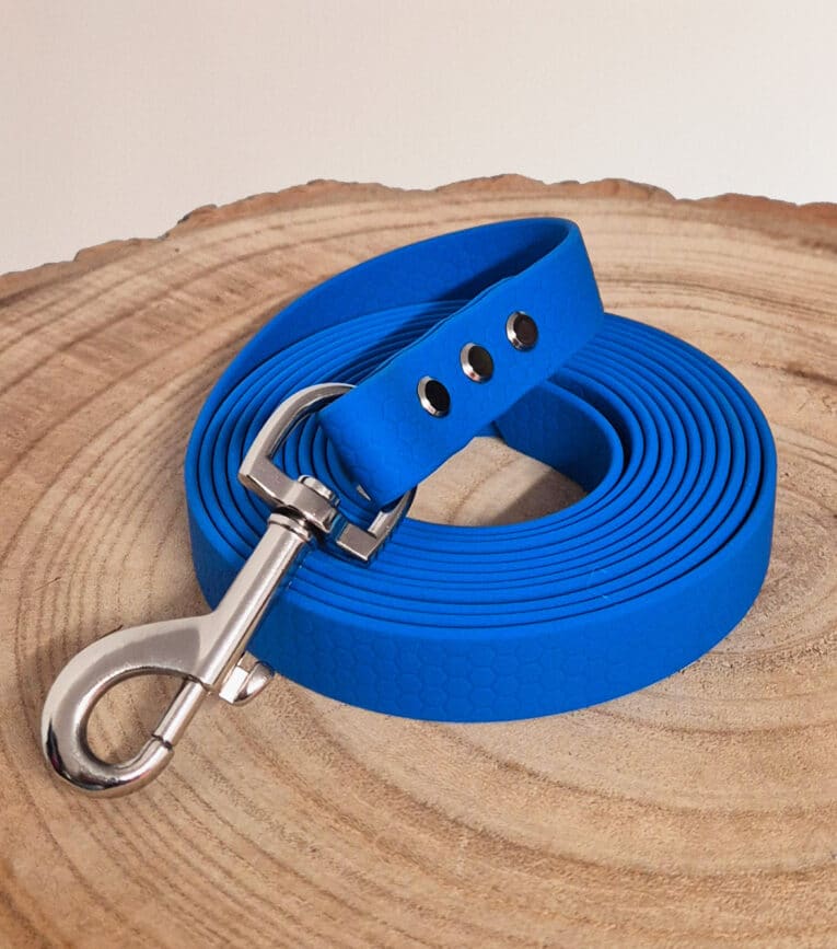 A rolled-up bright blue Waterproof Hexa Long-Line / Training Lead with a metal clasp lies on a round, light wood surface, featuring a subtle hexagonal pattern and three metal-reinforced holes.