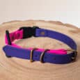 The Custom Quick-Snap Bungee Collar Strap in purple and pink features brass fittings and a black plastic buckle, shown on a round wooden surface.