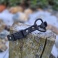 The Quick-Snap Bungee Collar, black with metal studs and a buckle, sits on a weathered tree stump outdoors amid snow patches, rocks, and green foliage.