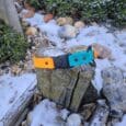 The Quick-Snap Bungee Collar, featuring a bright orange and teal design with an easy Quick-Snap buckle, sits on a weathered wooden post among snow, rocks, and greenery in an outdoor garden.
