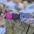 A Quick-Snap Bungee Collar, half pink and half grey, rests on a weathered tree stump outdoors with snow and blurred natural scenery in the background.