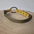 The Quick-Snap Bungee Collar, featuring olive green and yellow straps, metal rivets, and a black Quick-Snap ring, rests on a round light wooden surface against a plain background.