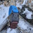 A Quick-Snap Bungee Collar, featuring blue and brown colours with a black buckle, rests on a snow-dusted wooden surface outdoors among rocks and plants.