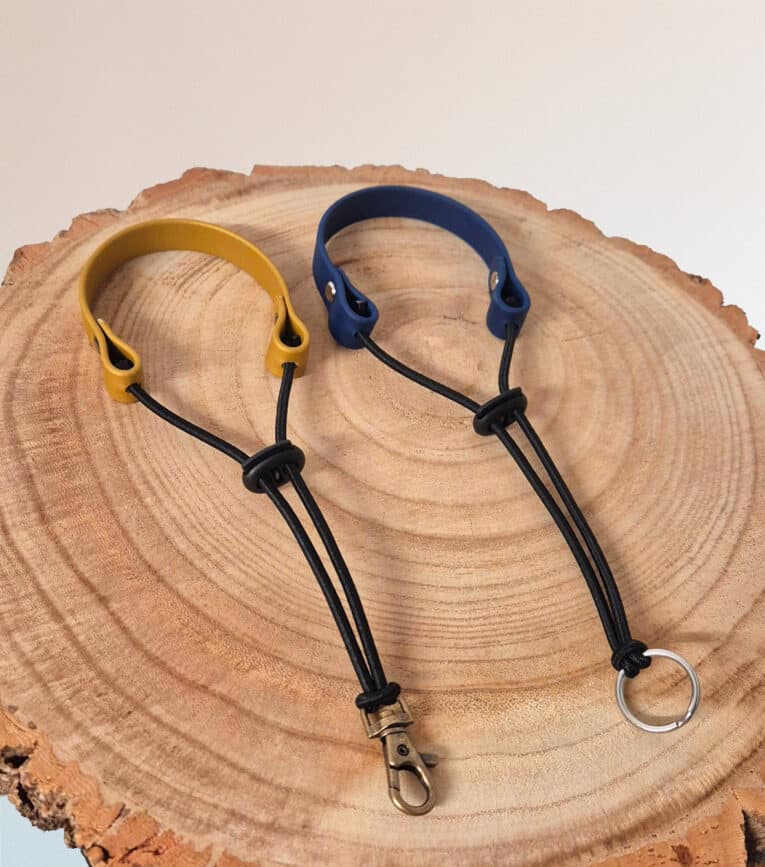 Two Super-grip Biothane Grab Tabs—one yellow with a metal clip, one blue with a metal ring—rest side by side on a round wooden slab. Perfect as customisable traffic handle leads.