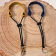 Two Super-grip Biothane Grab Tabs/Handles—one with a yellow loop and metal clasp, one with a blue loop and keyring—are displayed side by side on a round wooden surface, highlighting their customisable use as Traffic Handle Leads.
