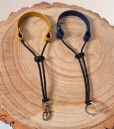 Two Super-grip Biothane Grab Tabs/Handles—one with a yellow loop and metal clasp, one with a blue loop and keyring—are displayed side by side on a round wooden surface, highlighting their customisable use as Traffic Handle Leads.