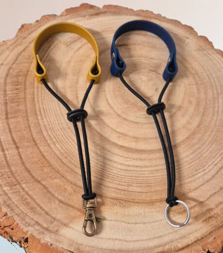 Two Super-grip Biothane Grab Tabs/Handles—one with a yellow loop and metal clasp, one with a blue loop and keyring—are displayed side by side on a round wooden surface, highlighting their customisable use as Traffic Handle Leads.