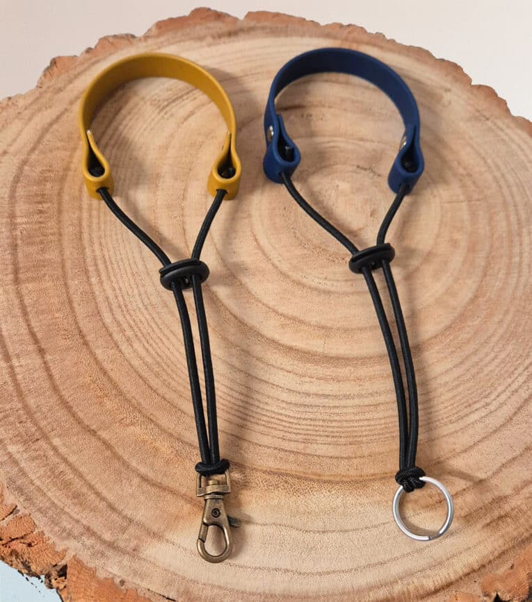 Two Super-grip Biothane Grab Tabs/Handles—one with a yellow loop and metal clasp, one with a blue loop and keyring—are displayed side by side on a round wooden surface, highlighting their customisable use as Traffic Handle Leads.