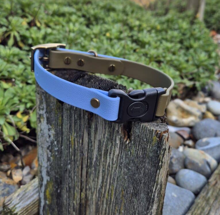 The Custom Quick-Snap Collar Strap in blue and olive green with brass fittings rests on a weathered wooden post, with smooth stones and green foliage softly blurred in the background.