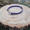 The Custom Snap Tag/Slip Collar in purple is shown on a round wooden tree slice, featuring a metal clasp and D-ring, surrounded by grass and small stones.