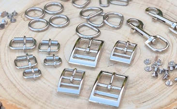 Various shiny silver metal hardware pieces, including best dog lead fittings such as buckles, D-rings, clasps, and screws, are arranged neatly on a light-coloured wooden surface—ideal for crafting or assembling quality dog collars.