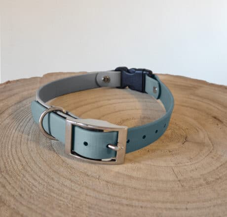 The Custom Quick-Snap Bungee Collar (Copy) features a teal and grey design with a silver buckle and D-ring, shown on a round wooden surface with visible tree rings.