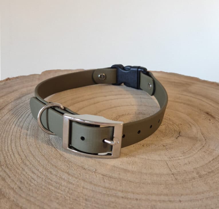 The Custom Quick-Snap Bungee Collar (Copy) in olive green, featuring a silver buckle and metal D-ring, is shown coiled on a round, textured wooden surface with visible tree rings.
