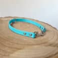 A turquoise pet collar with metal rivets and a clasp is displayed on a round, wooden surface with visible tree rings. The background is neutral and unfocused.