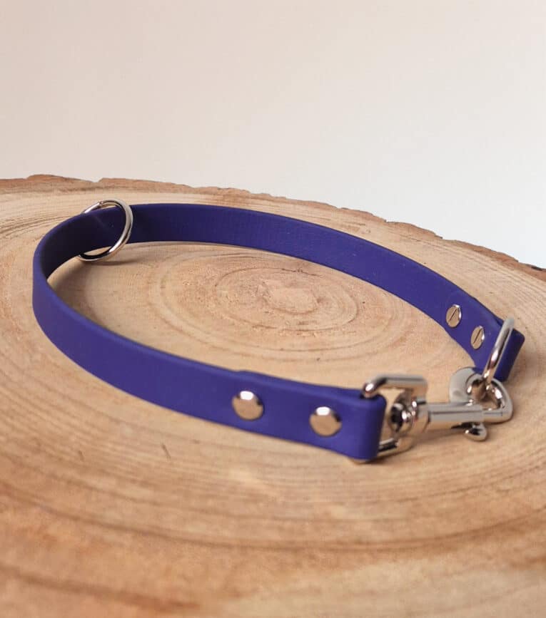 A purple dog collar with metal rivets, a D-ring, and a clasp is displayed on a round, light-coloured wooden surface.