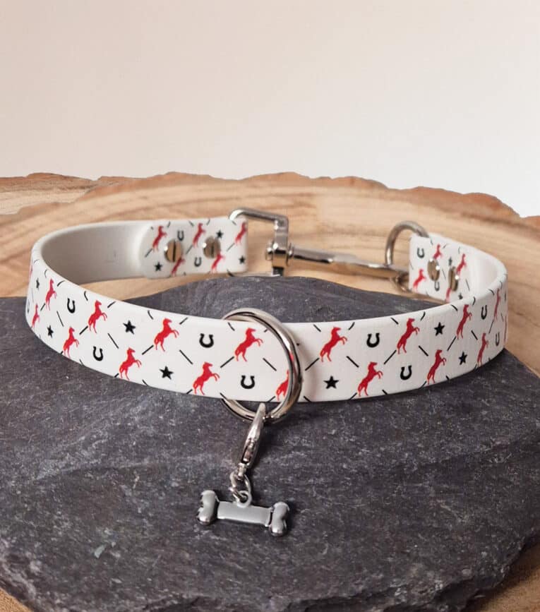 The Custom Snap Tag - Slip Collar is a white, quick-snap customisable dog collar with red horse, black star, and horseshoe patterns, plus a small silver bone charm. Displayed on grey stone with a light background.