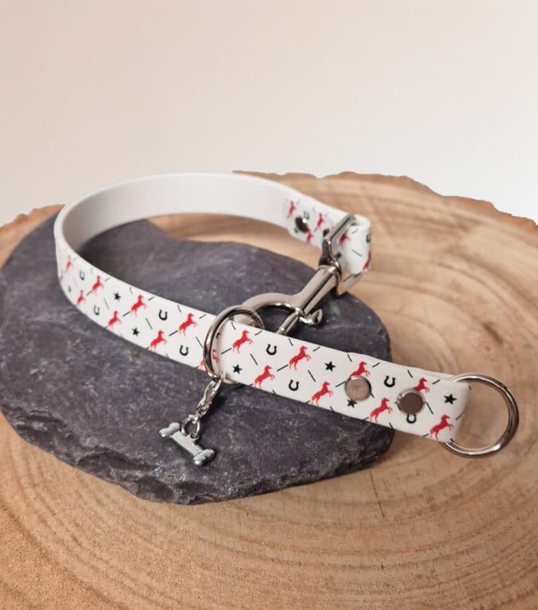 The Custom Snap Tag - Slip Collar features a white design with red horses and black horseshoes, metal fittings, and a small silver bone charm, shown on dark stone set on wood.