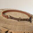 A brown and tan leather dog collar with metal rings and rivets is displayed on a wooden tree stump slice. The collar is arranged in a circular shape with a clasp visible on the right side.