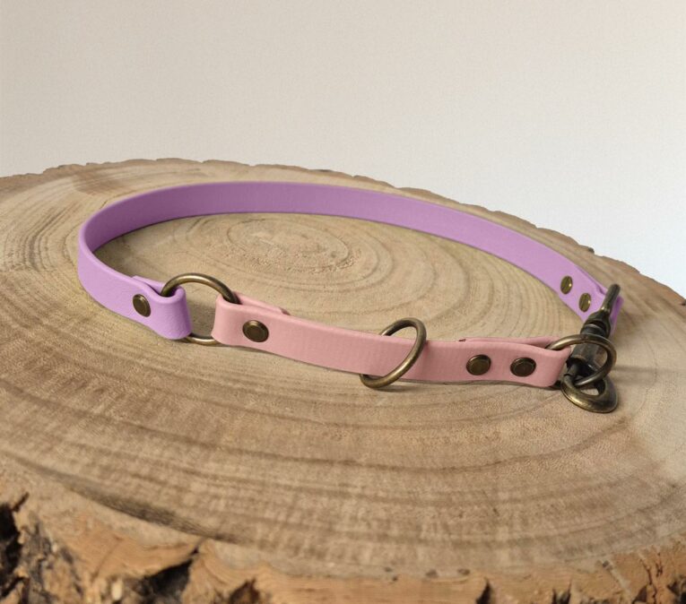 A two-tone dog collar in pastel purple and pink with brass fittings is displayed on a round, wooden surface with visible tree rings.