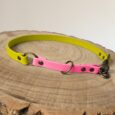A dog collar with a neon yellow band and a pink section, made of synthetic material with metal fittings, is displayed on a round wooden surface.