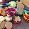 Several rolls of colourful paper or ribbon, secured with brown tape, are arranged on a cutting mat on a wooden table. The bright colours resemble the collar colour code often used for dog leads to indicate different temperaments or needs.