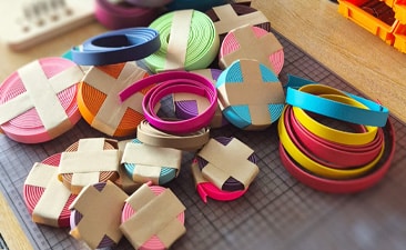 Several rolls of colourful paper or ribbon, secured with brown tape, are arranged on a cutting mat on a wooden table. The bright colours resemble the collar colour code often used for dog leads to indicate different temperaments or needs.