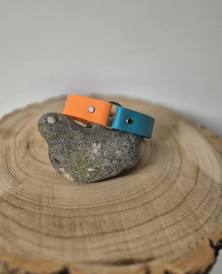 A grey bird-shaped stone sits on a wooden slab, adorned with orange and teal Centre O-Ring Collars (Gundog Collars). The background is plain and blurred.