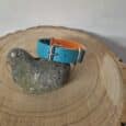 A blue and orange strap with a silver buckle, styled like the Centre O-Ring Collar / Gundog Collar, is shown on a bird-shaped stone atop a round wooden surface with visible tree rings.