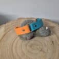 A Centre O-Ring Collar / Gundog Collar in two-tone orange and teal with a metal buckle is shown on a smooth grey stone placed atop a wooden surface featuring visible tree rings.