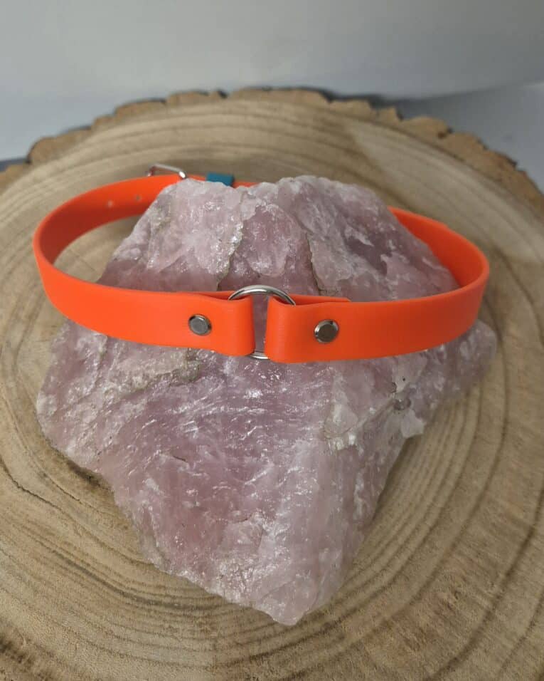The Centre O-Ring Collar / Gundog Collar, featuring metal fittings and an orange design, is shown draped over a large pink crystal on a round wooden surface.