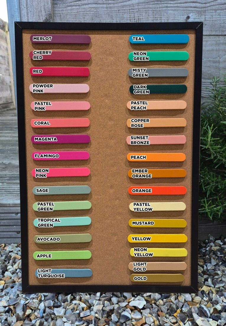 A corkboard displays paint colour samples in rows—merlot, cherry red, teal, sage, peach, and gold—resting on gravel by a plant and fence, echoing the vibrant tones of the Centre O-Ring Collar / Gundog Collar.