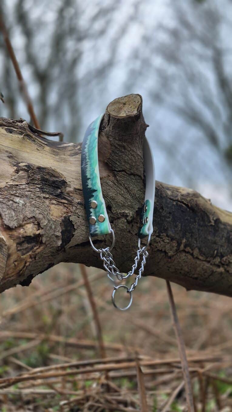 A Martingale / Half-Check Collar in a green and black pattern with a metal chain and ring is draped over a fallen tree branch outdoors, with blurred trees and grass in the background.