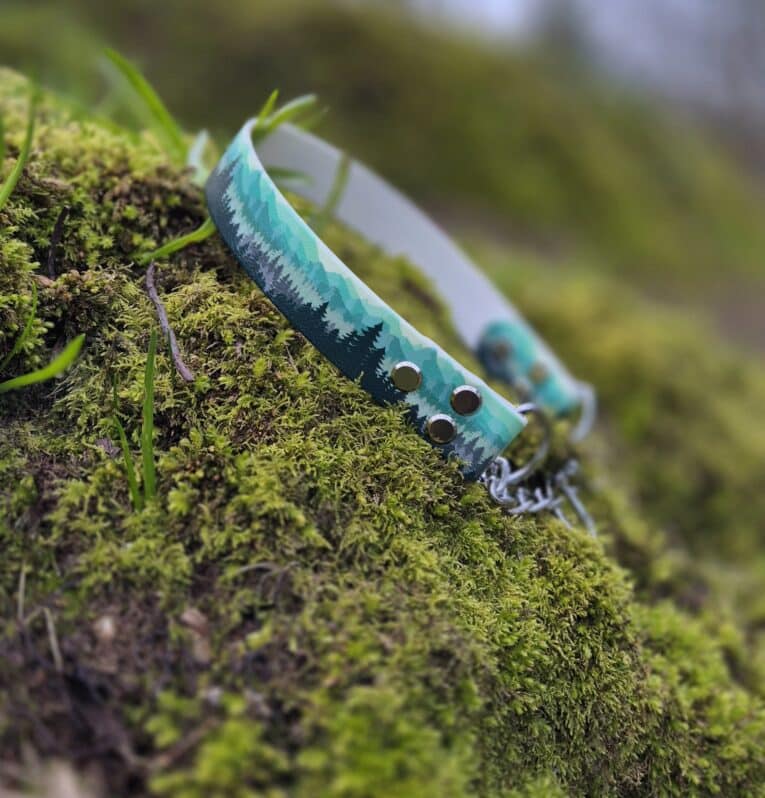 A Martingale / Half-Check Collar with a forest tree pattern rests on vibrant green moss outdoors, featuring metal eyelets and a clasp. The blurred background is filled with lush greenery.
