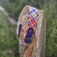 A vibrant check Martingale / Half-Check Collar with purple leather and metal chain accents rests on a weathered wooden post, set against a backdrop of blurred green foliage.