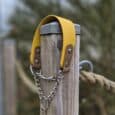 A Martingale / Half-Check Collar in yellow and brown with a metal chain section hangs on a weathered wooden post outdoors, alongside a thick rope and blurred greenery in the background.