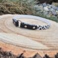 The Tag Collars dog collar, featuring a black-and-white cow print and metal ring, rests on a cut wooden stump outdoors with grass and rocks in the background.