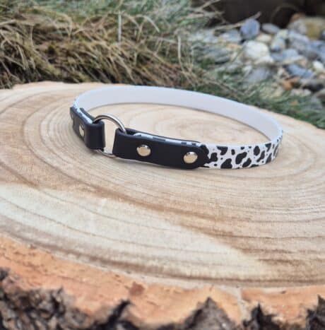 The Tag Collars dog collar, featuring a black-and-white cow print and metal ring, rests on a cut wooden stump outdoors with grass and rocks in the background.