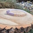 The Tag Collars pet collar, featuring white and lavender colours with colourful flower designs, is displayed on a tree stump outdoors amidst grass and rocks.