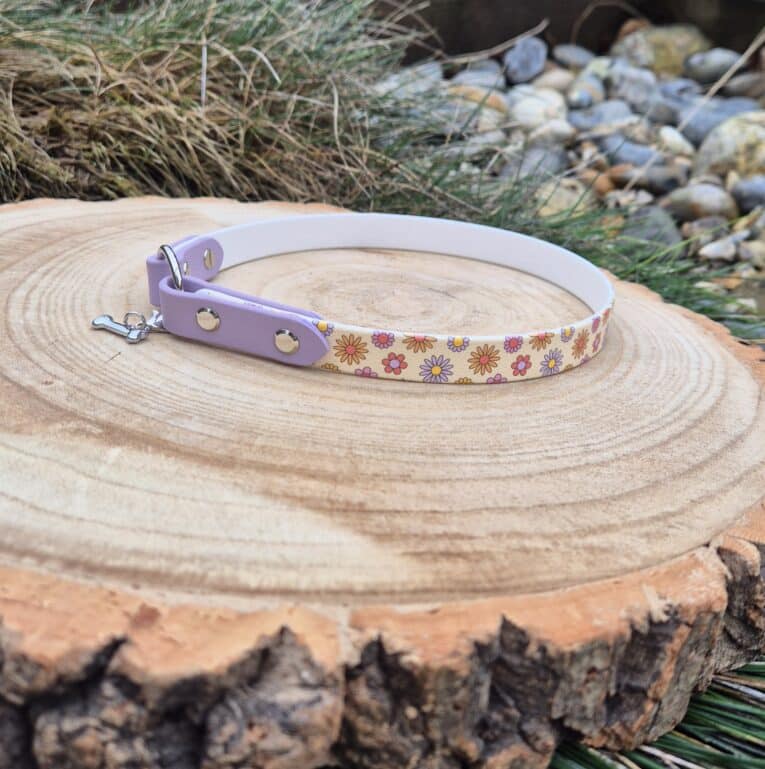 The Tag Collars pet collar, featuring white and lavender colours with colourful flower designs, is displayed on a tree stump outdoors amidst grass and rocks.
